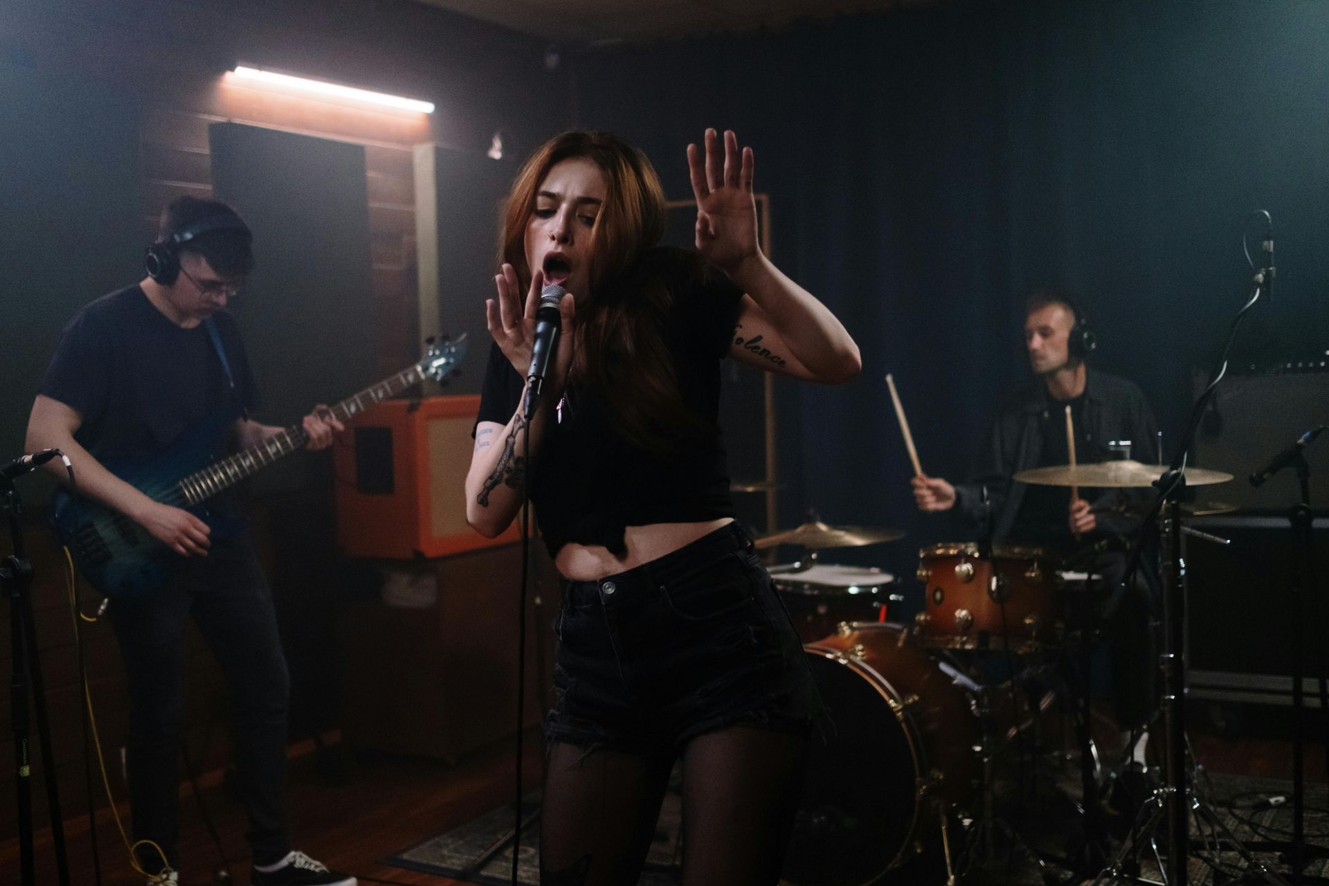 Band performing in a dim rehearsal room, singer at center with guitarist and drummer behind.