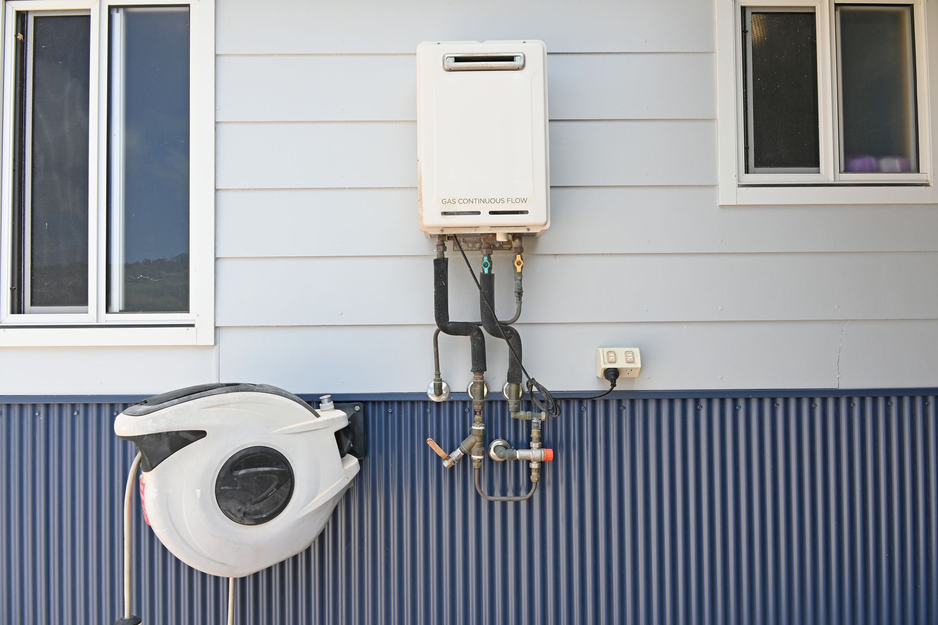 Wall-mounted gas water heater with pipes on a house exterior, above a coiled white hose.