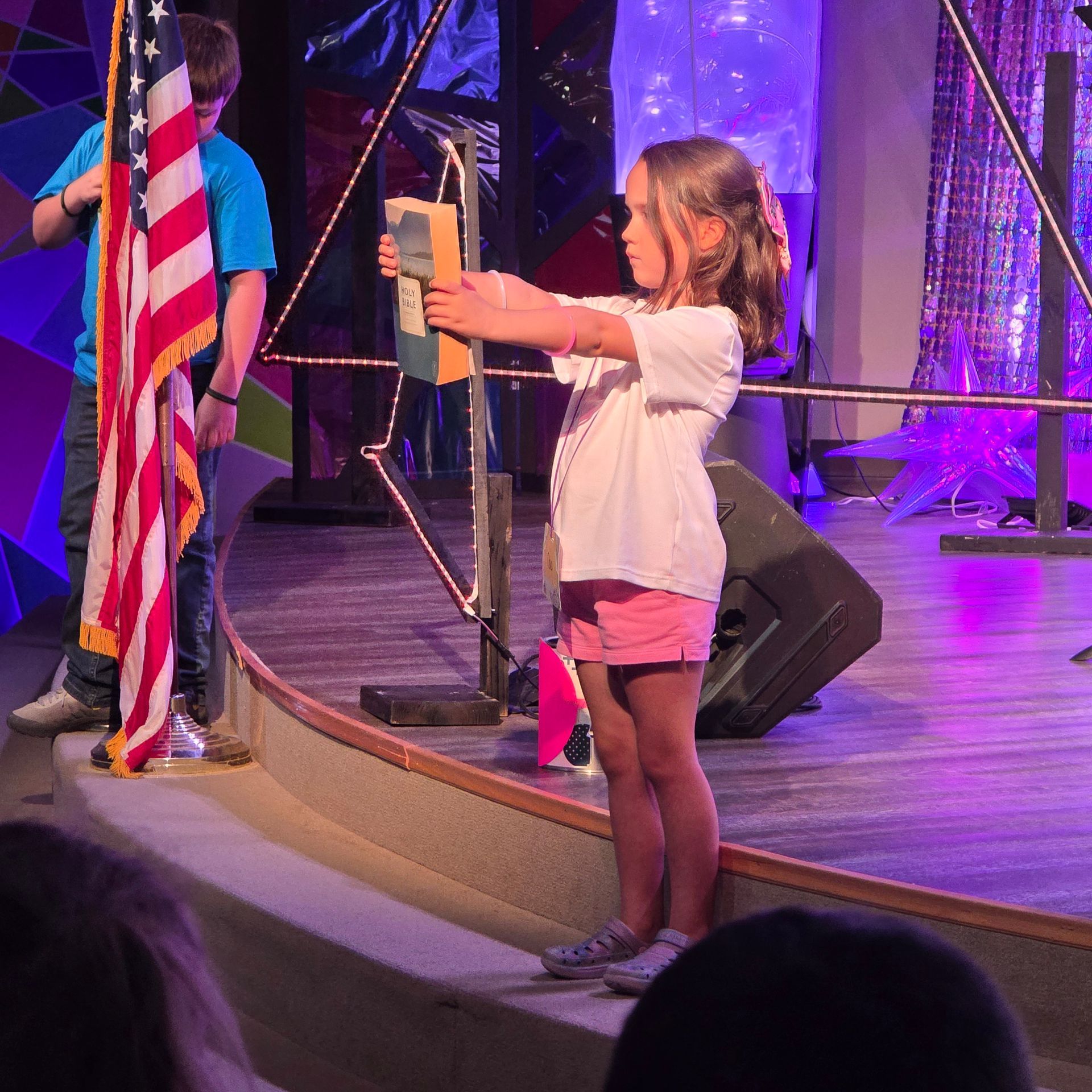 Child holding up a certificate on a stage beside a U.S. flag and microphone stand.