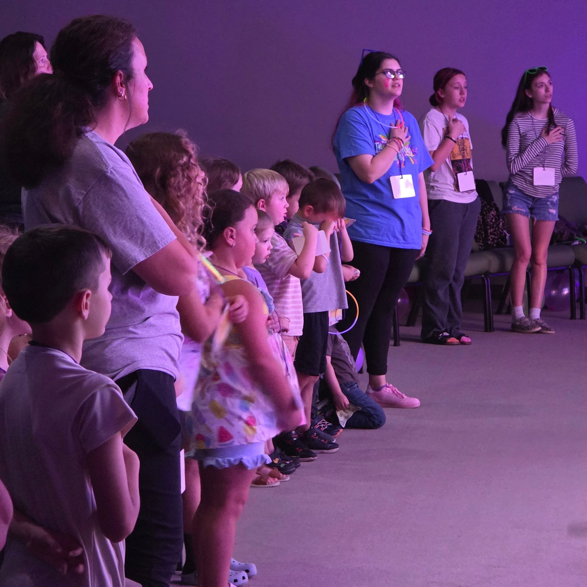Children and adults standing in a purple-lit event space, watching something off to the left.