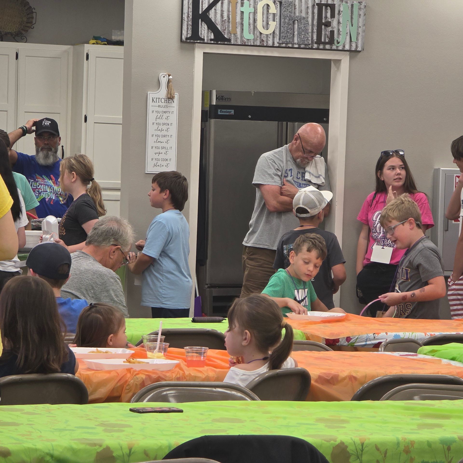 Children and adults gathered around orange and green tables in a community room with a KITCHEN sign.