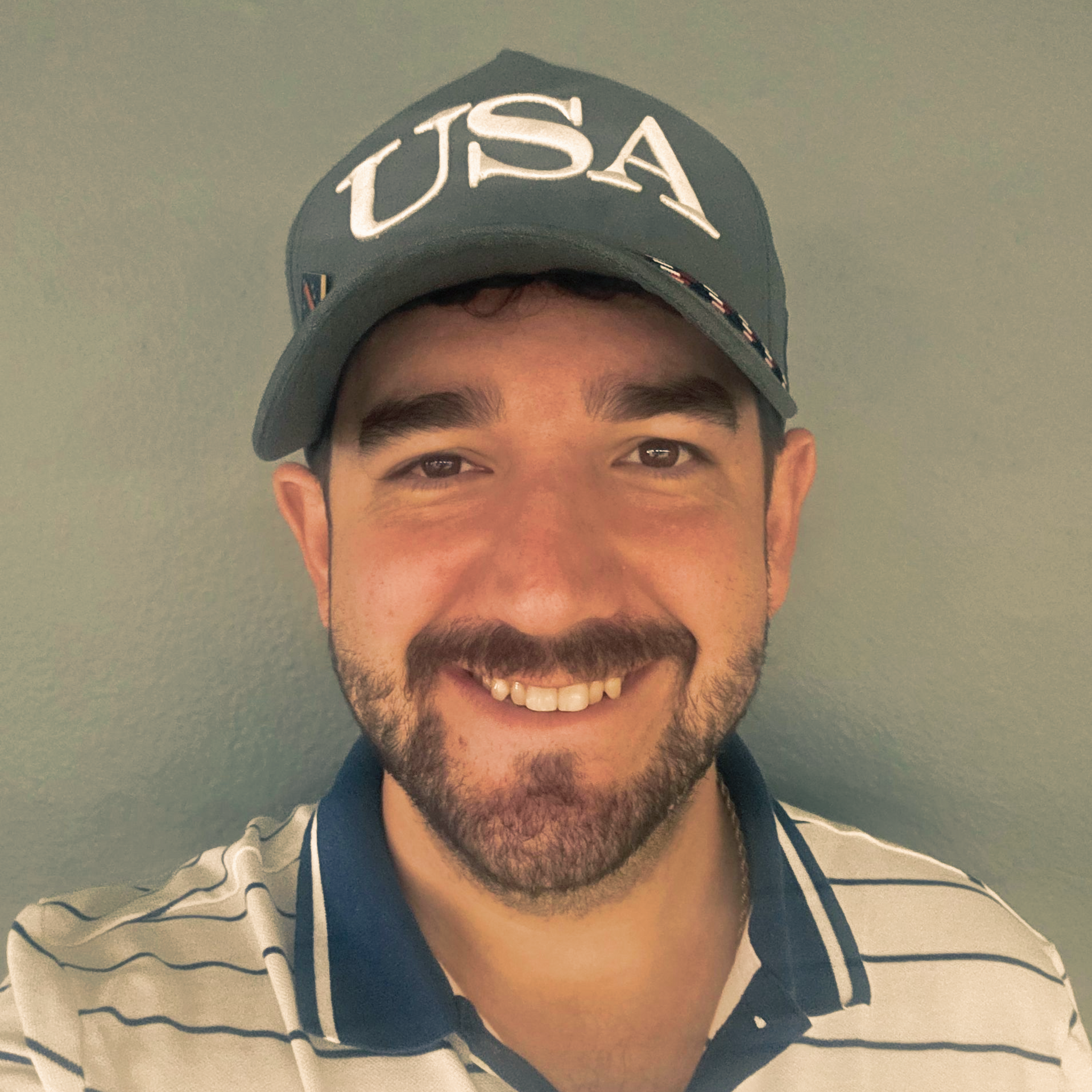 Smiling man wearing a USA cap and striped polo shirt against a plain wall