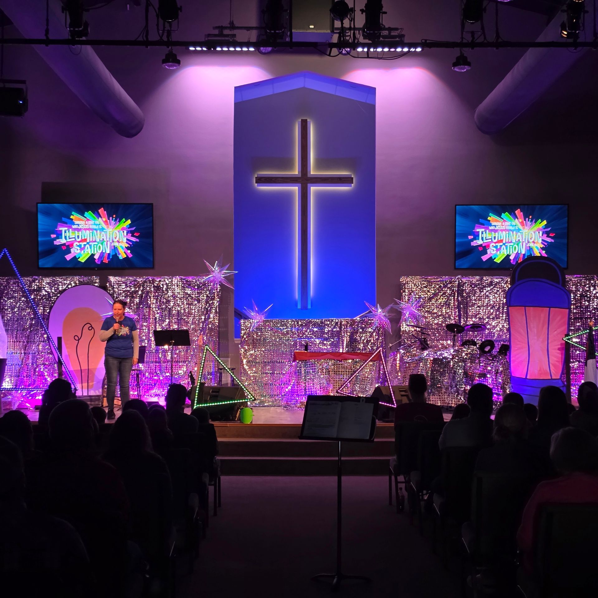 Church stage with glowing cross, purple lighting, musicians, and audience silhouettes