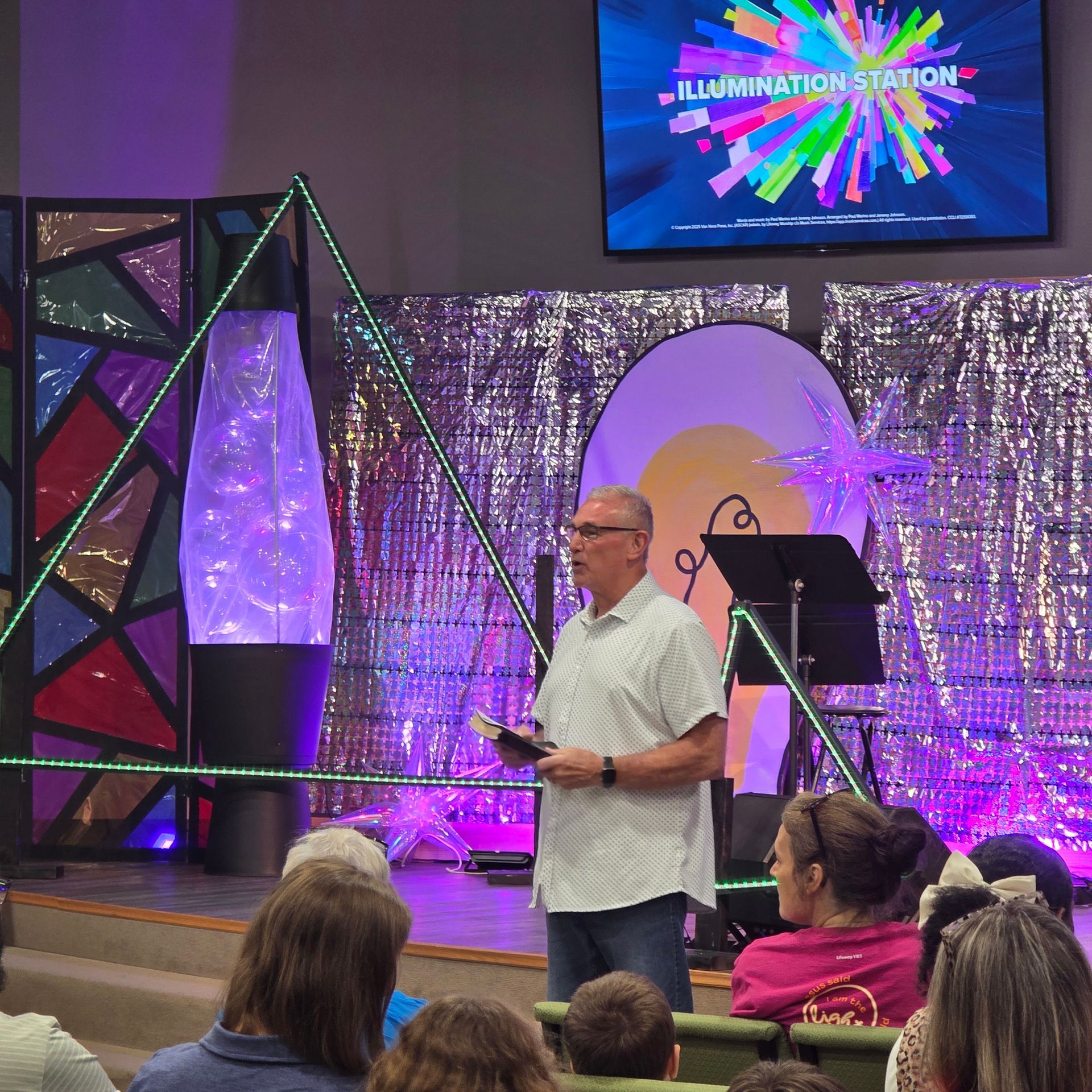 Man speaking on stage in front of a purple-lit backdrop, with audience seated and a screen above.