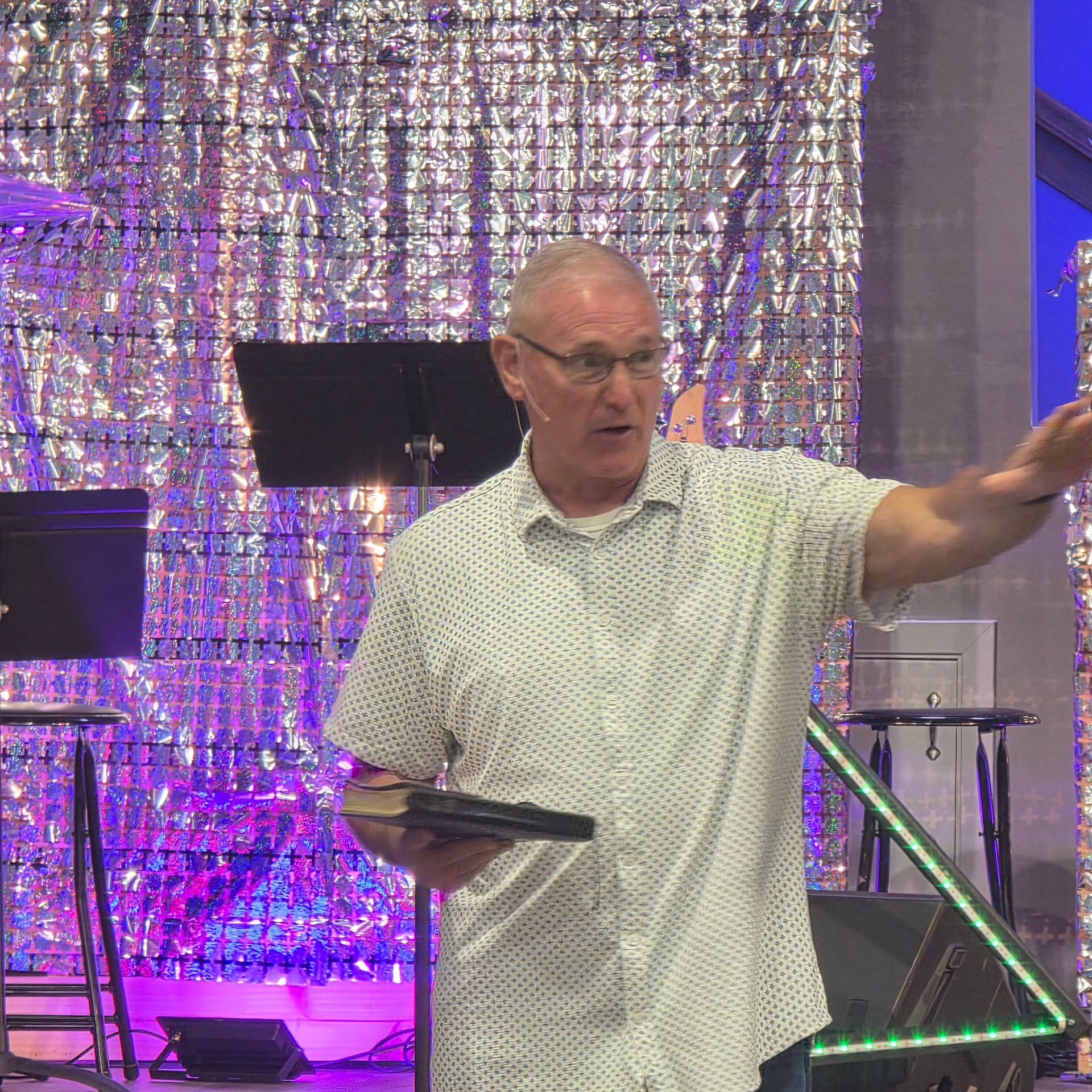 Man speaking on stage, holding a tablet and gesturing beside purple-lit backdrop and music stands