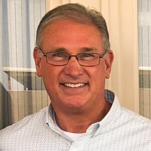 Smiling man in glasses and a light blue shirt standing indoors by a window