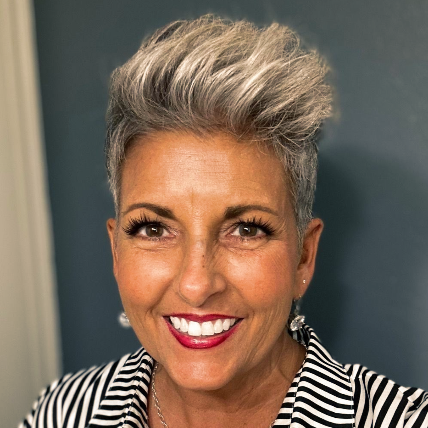 Smiling person with short silver hair wearing a black-and-white striped shirt against a dark background