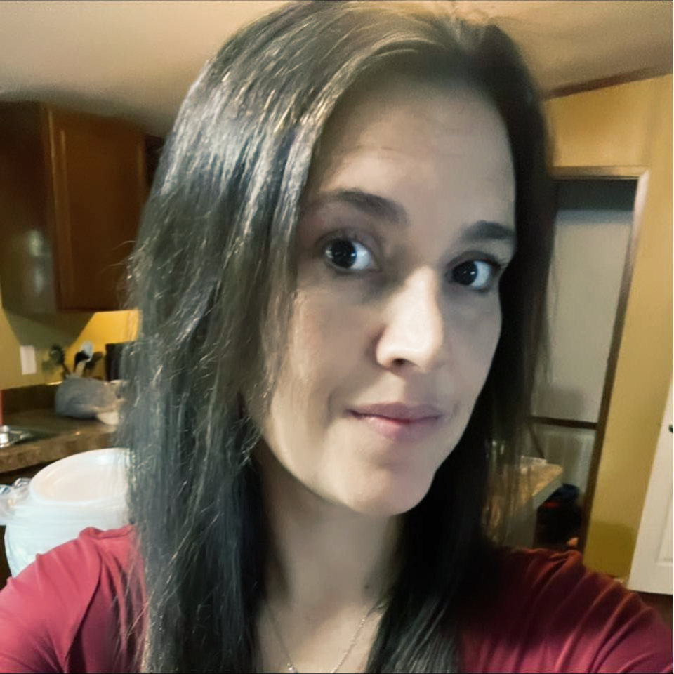 Close-up selfie in a kitchen, showing a person with long brown hair and a slight smile.