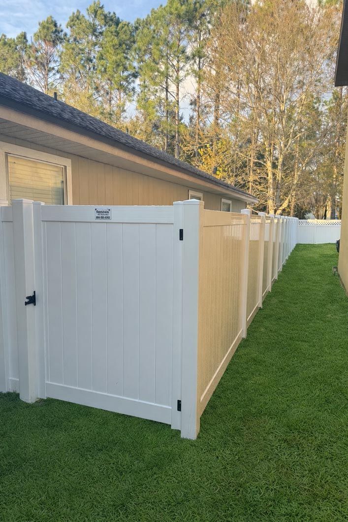 White vinyl fence and gate along a narrow grassy side yard beside a beige house and trees