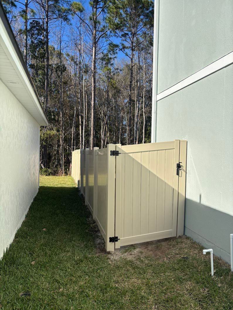 Narrow grassy side yard with a beige gate between two light-colored houses, backed by trees and blue sky