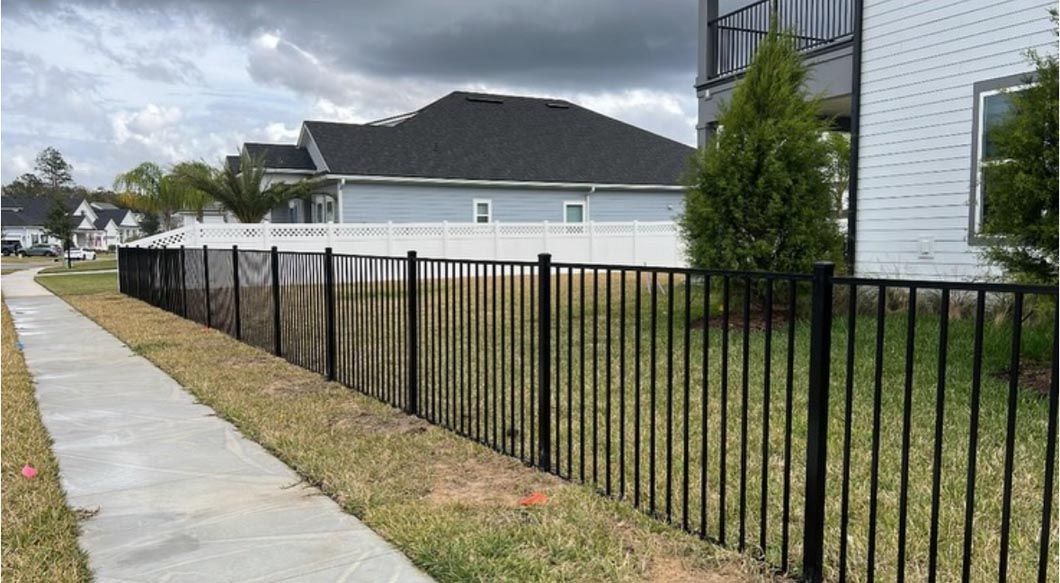Sidewalk beside a black metal fence and grassy yard, with a light blue house under cloudy skies.