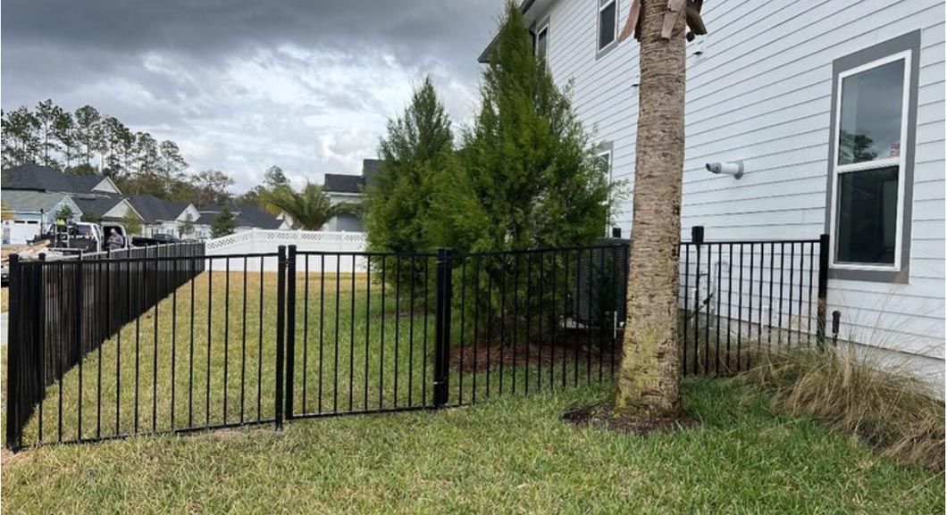 Black metal fence beside a white house and grass yard with a tall tree and overcast sky