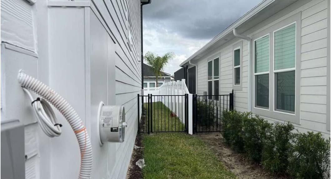Narrow side yard between two white houses with utility equipment, grass, shrubs, and a gate in back