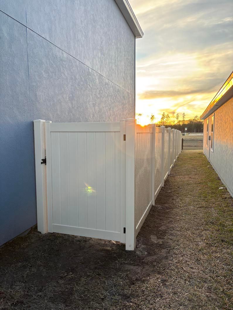 White fence gate beside a blue building at sunset, with a narrow gravel path along the side yard