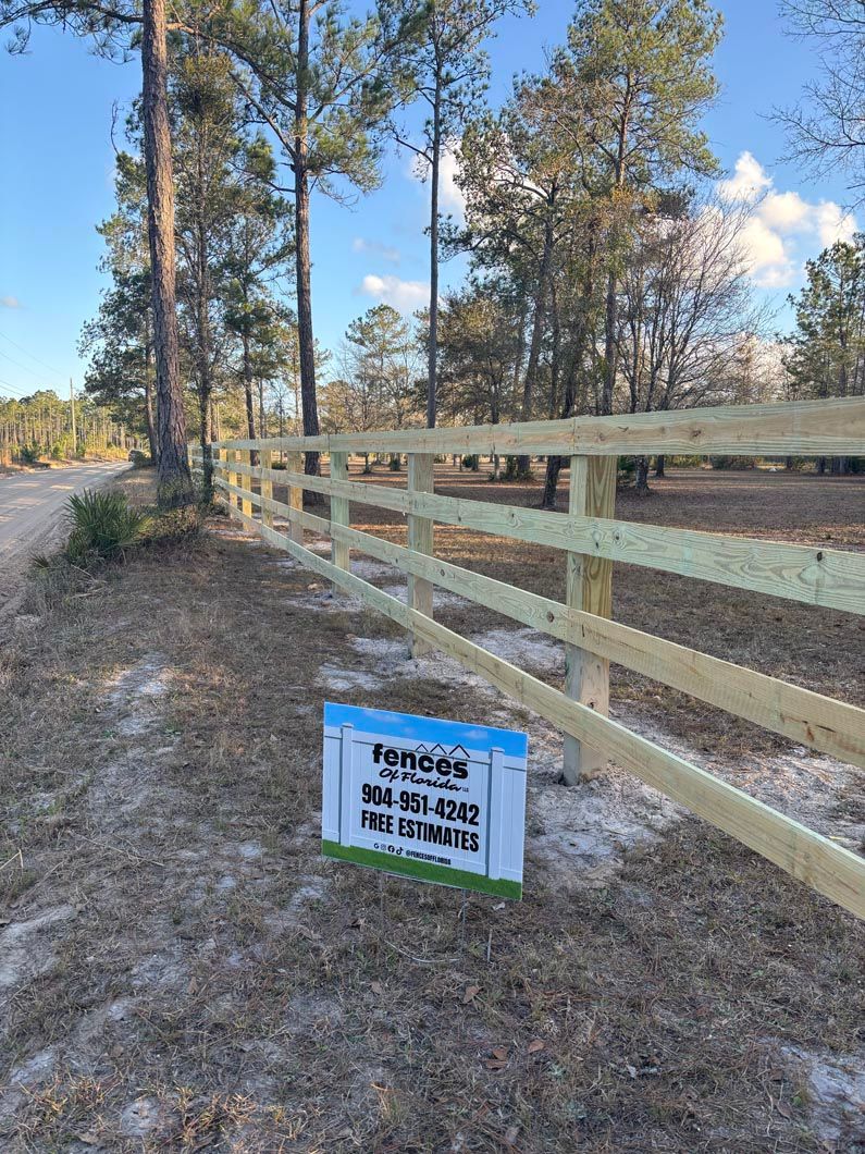 Wooden fence along a dirt road in a wooded area, with a blue real estate sign in the foreground.