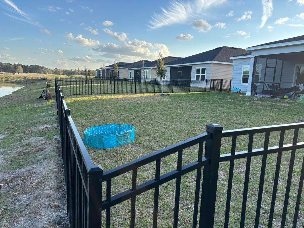 Backyard with black fence, small blue kiddie pool, and row of houses under a cloudy sky