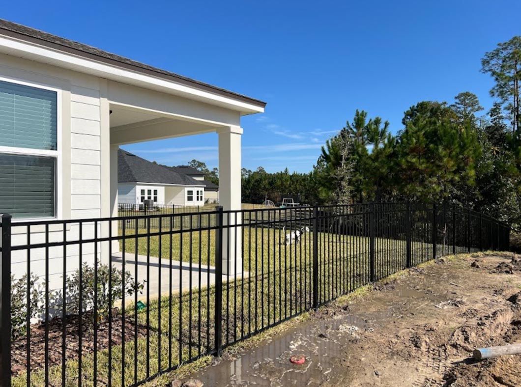 Black metal fence beside a white house porch and dirt yard under a clear blue sky