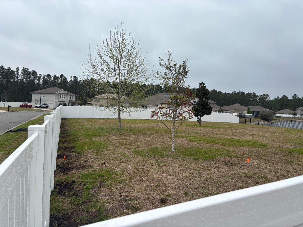 White fence borders a yard with small trees and a pond under a cloudy sky
