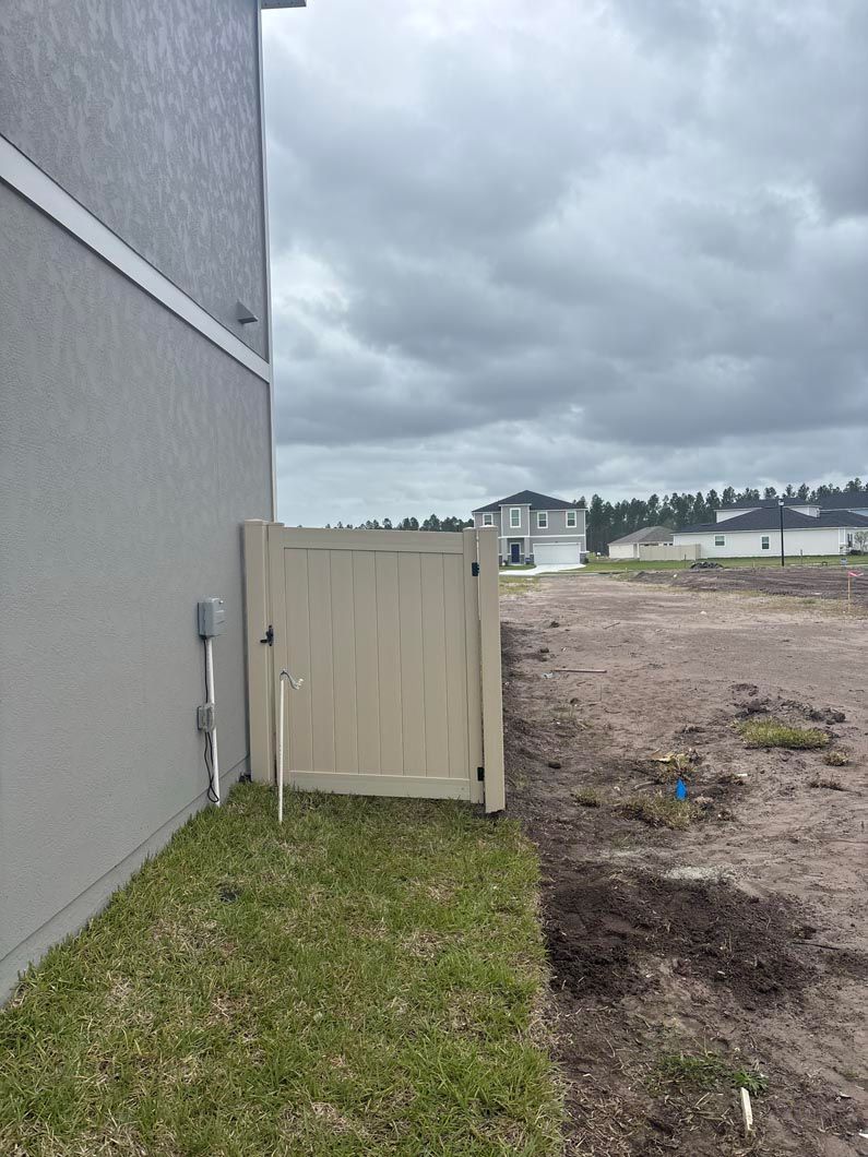 Open beige gate beside a gray house, overlooking a muddy vacant lot under cloudy skies.