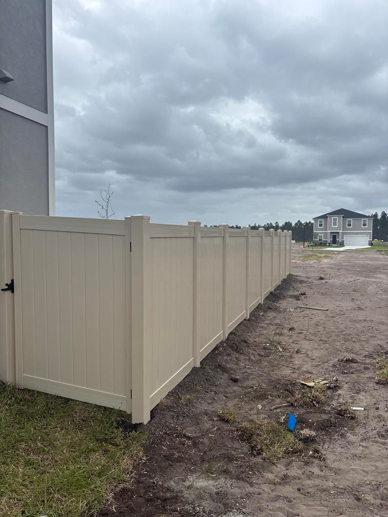 Beige vinyl fence along a muddy construction lot under cloudy skies, with houses in the distance.