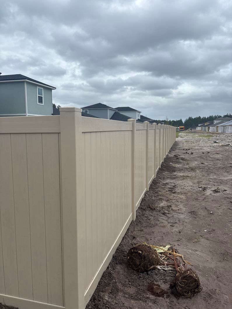 Beige vinyl fence along a dirt lot under a cloudy sky, with houses in the distance.