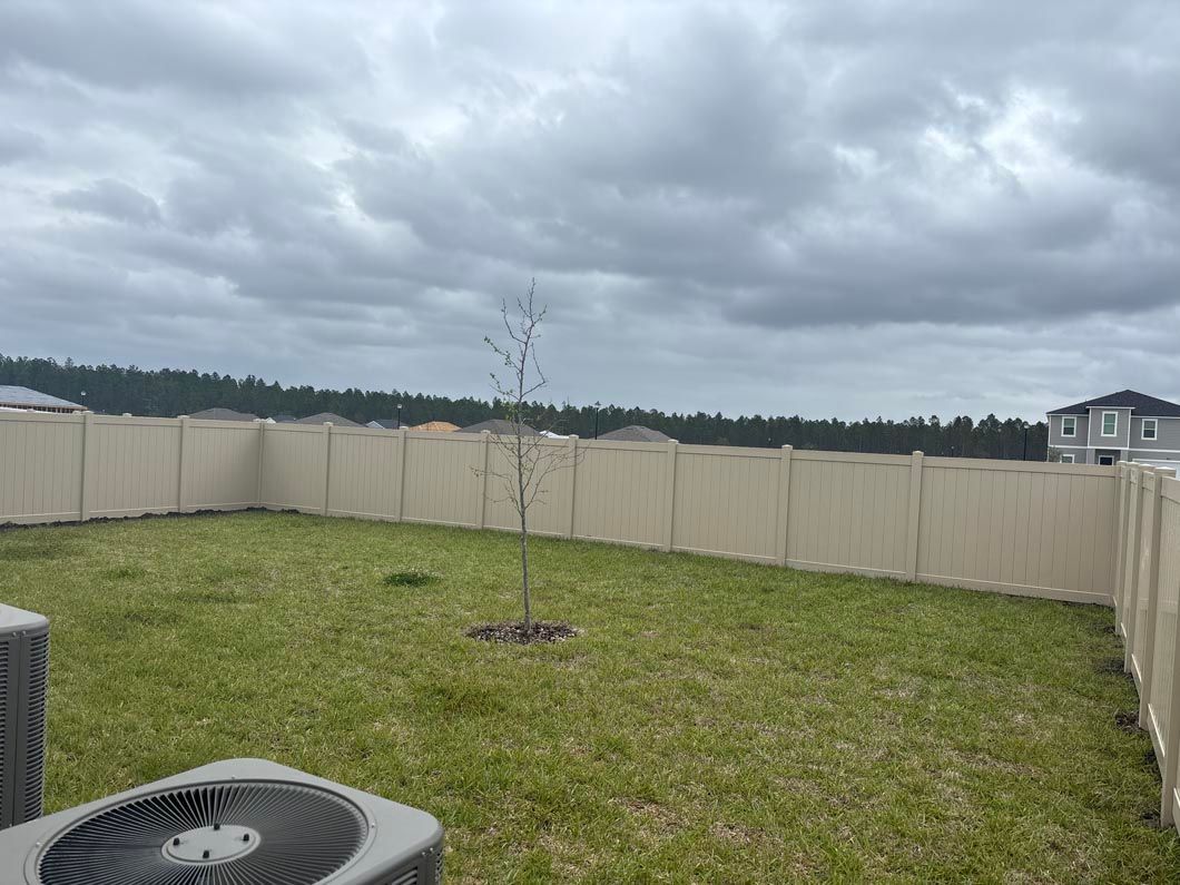 Grassy backyard with a young tree, beige fence, and cloudy sky above an AC unit.