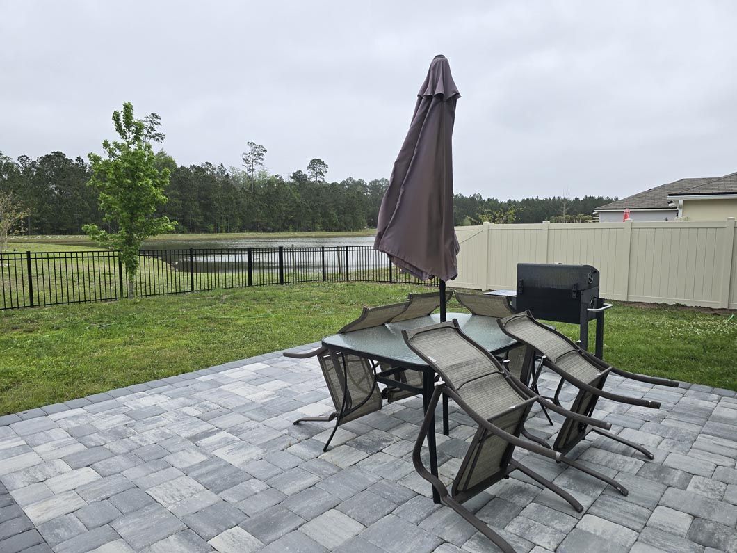 Patio with glass table and chairs under a closed umbrella, beside a grassy yard and pond.