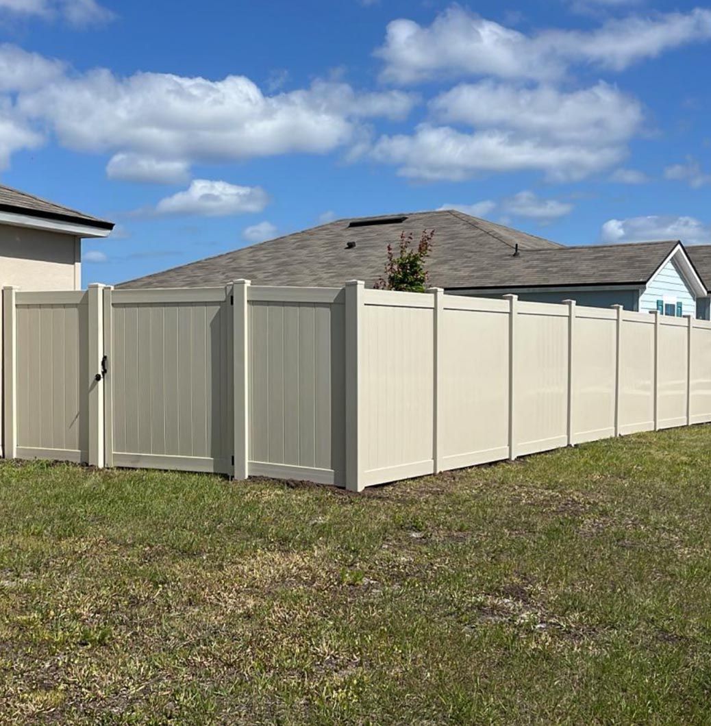 A long beige vinyl fence enclosing a backyard under a blue sky with clouds.