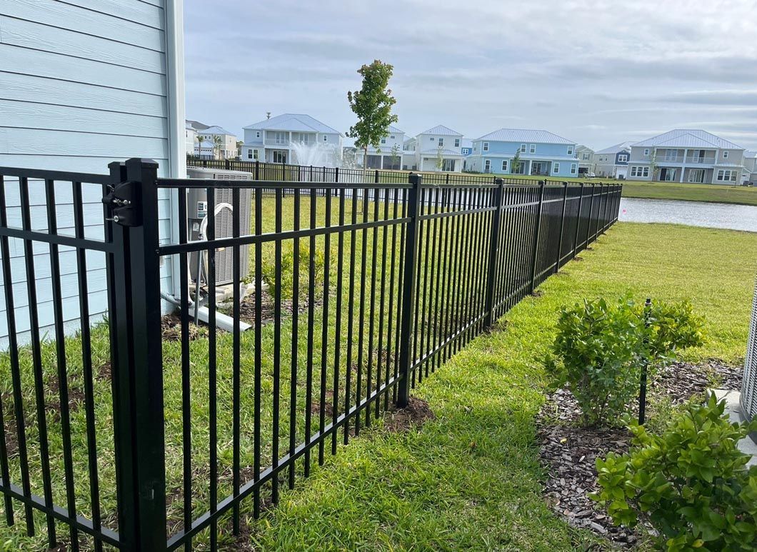Black metal fence beside a white house, overlooking a grassy yard and water with houses across the lake
