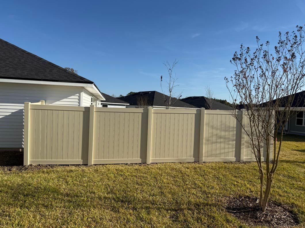 Beige vinyl privacy fence in a suburban backyard with a tree and grass under a blue sky