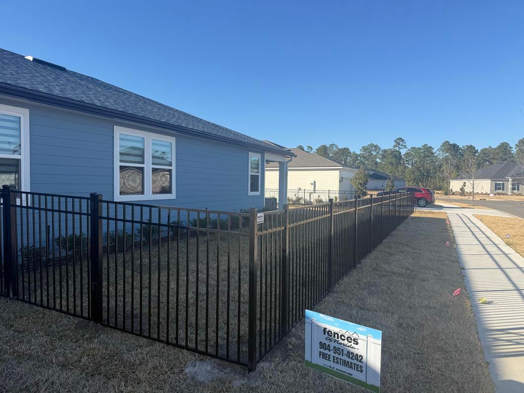 Black metal fence beside a blue house in a gravel yard with a small sign and clear sky.