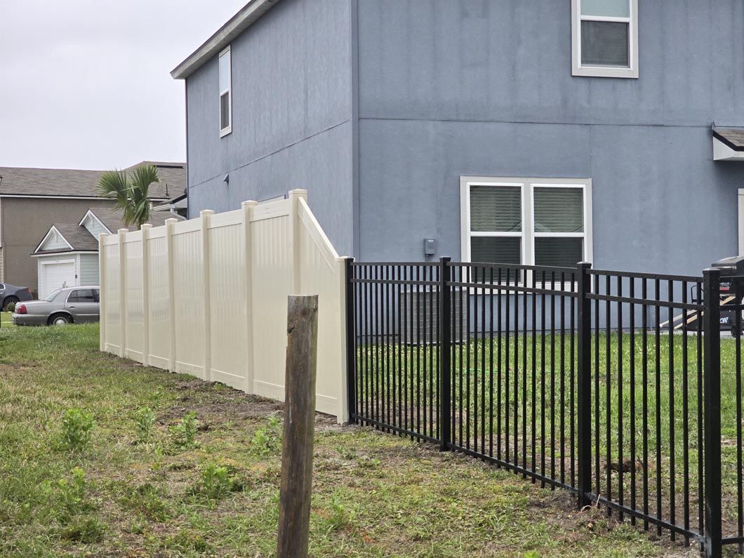 Backyard with beige privacy fence, black metal fence, and blue house beside a grassy lot