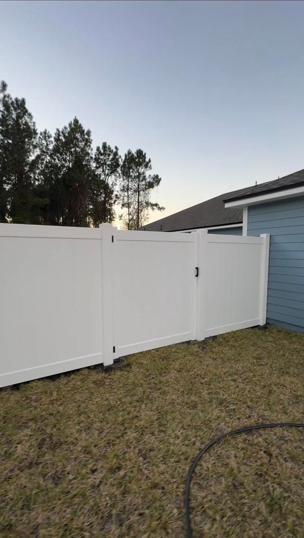 White vinyl fence in a grassy yard at sunset, with trees and a blue house in the background
