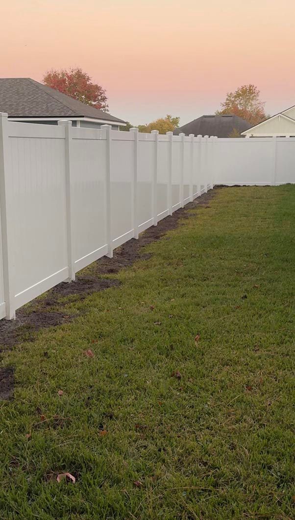 White vinyl fence lining a grassy backyard at sunset, with neighboring roofs in the background