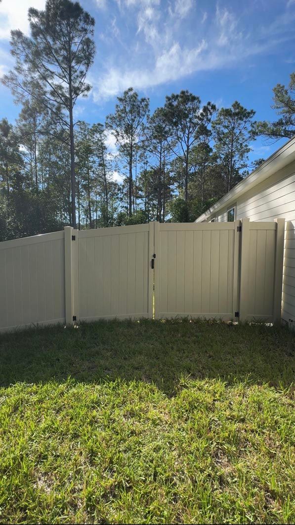 White backyard fence with a closed gate, green grass, and tall pine trees under a blue sky.