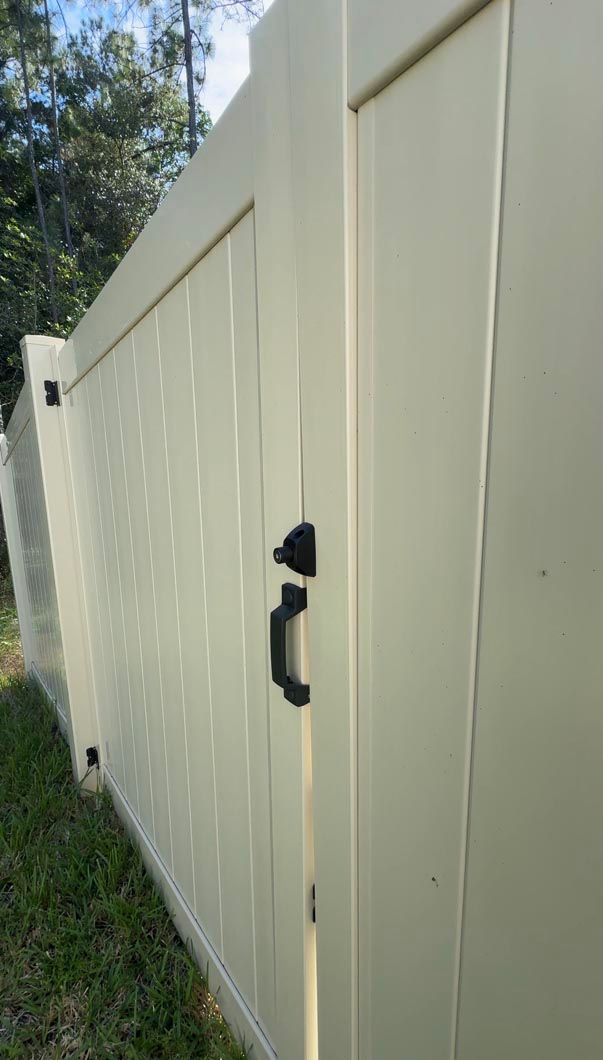 Beige vinyl fence with a black latch and gate in a grassy yard under trees.