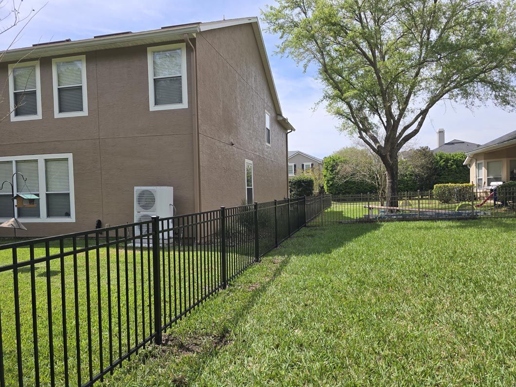 Brick house beside black fence and green lawn, with a large tree in the yard.