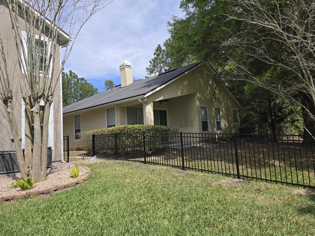 Single-story house with a sloped roof, black fence, and trees in a grassy yard.