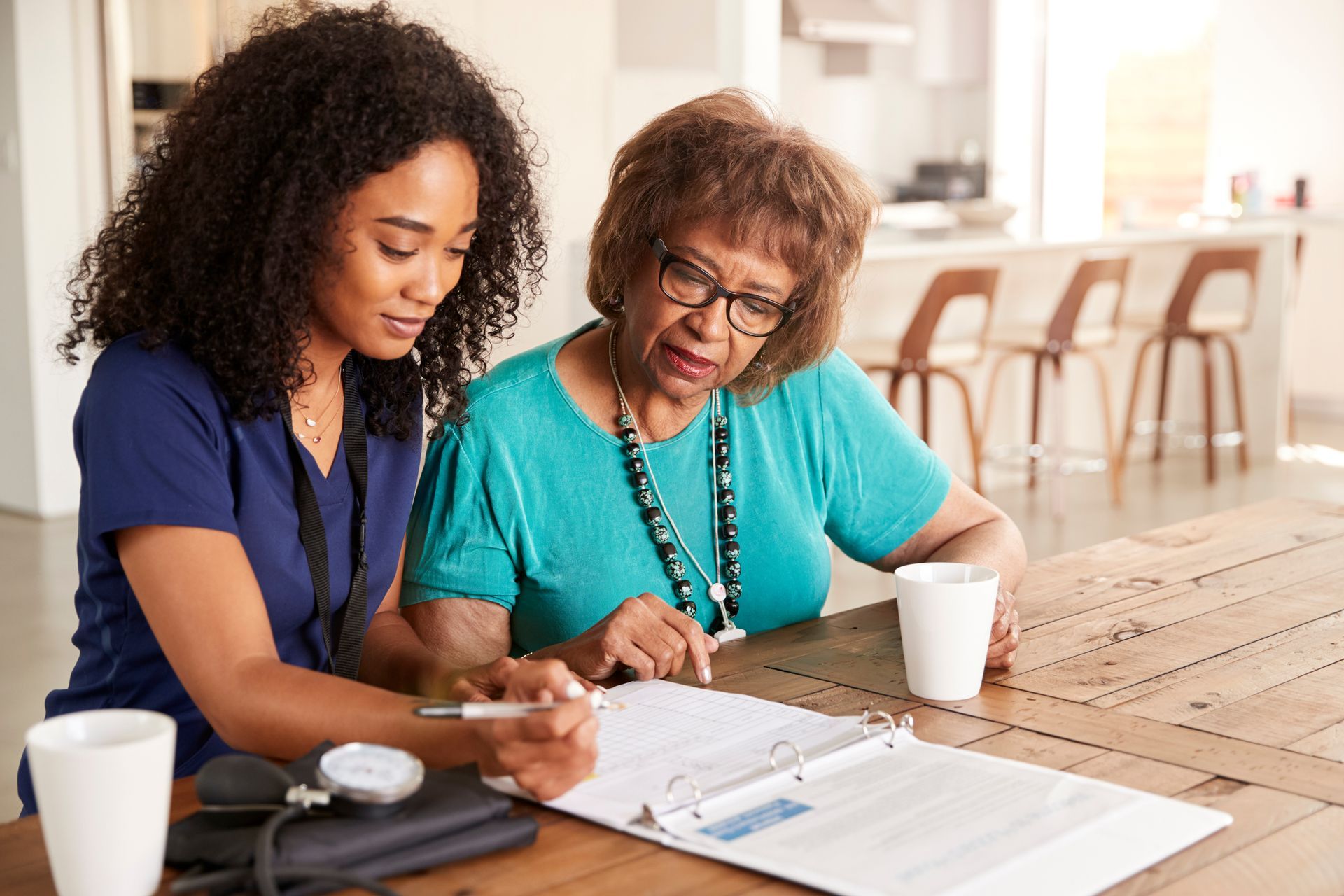 A healthcare professional sits at a table with an older person, reviewing documents together in a brightly lit home.