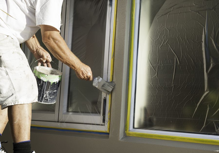 Person painting a window frame, holding paint can and brush. Windows are covered with plastic.