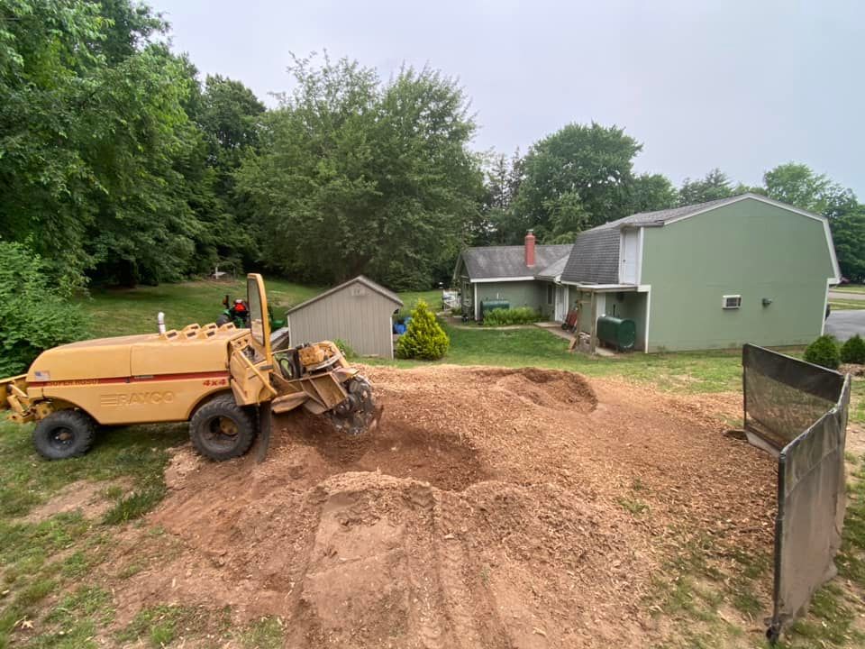 A yellow stump grinder working on a pile of dirt in a grassy yard in front of a green two-story house.