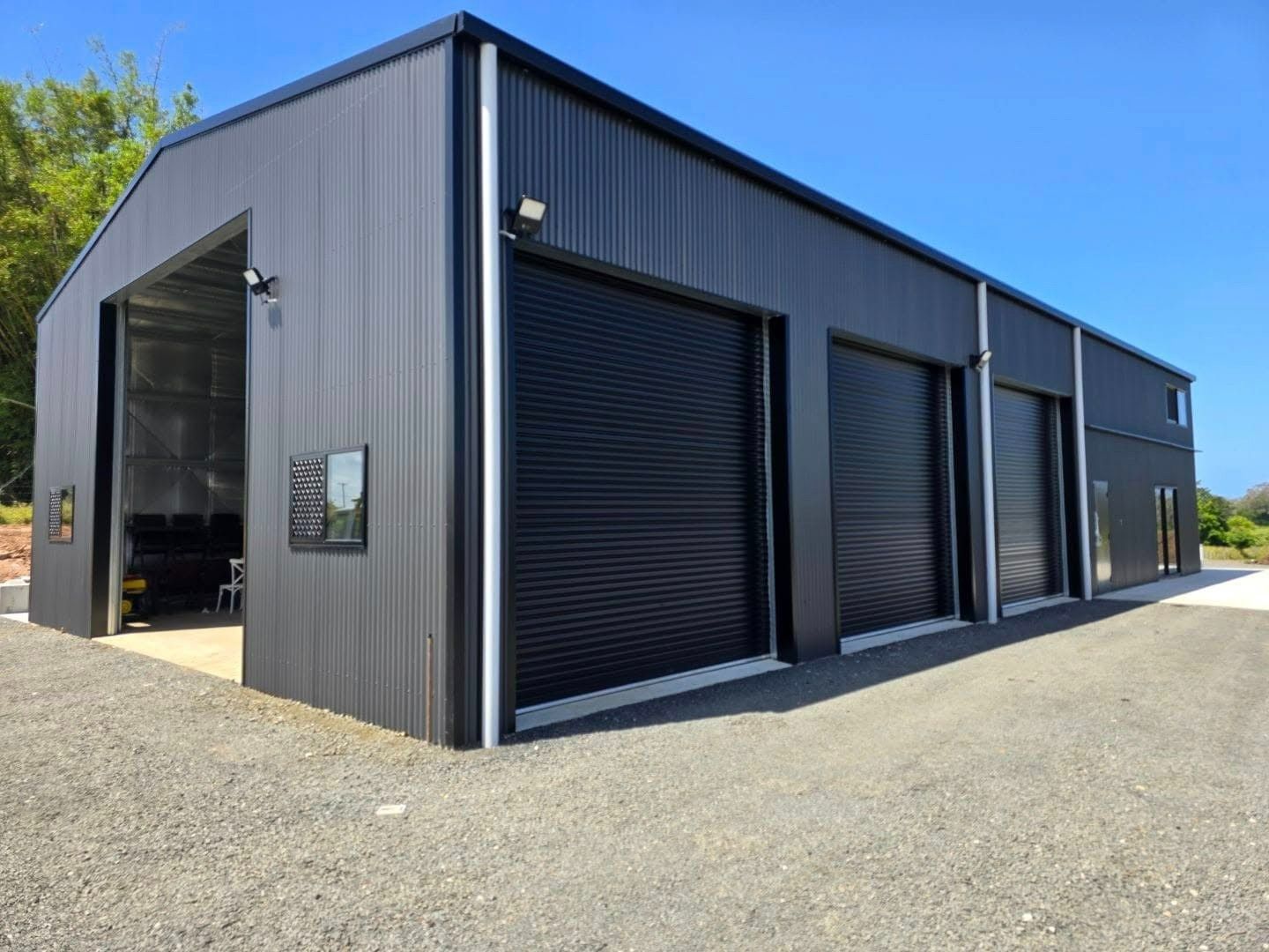 Black Metal Industrial Building With Three Garage Doors and an Open Bay. the Structure Sits on Gravel, Under a Bright Blue Sky — North Coast Sheds & Garages in Coraki, NSW