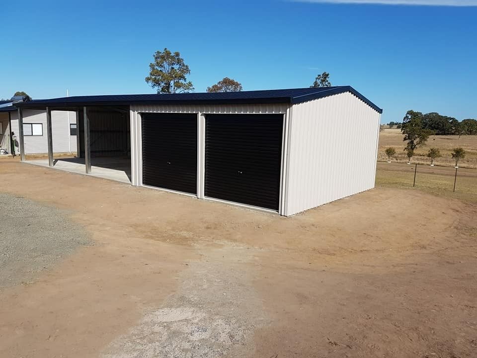 A Light-colored Metal Shed With Black Garage Doors and a Carport, Set on a Dirt Driveway Under a Blue Sky — North Coast Sheds & Garages in Coraki, NSW