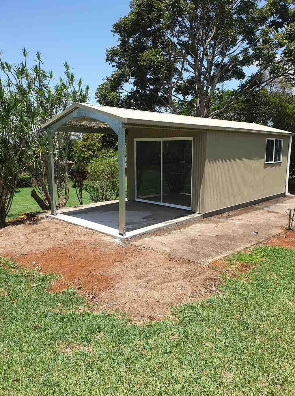 A Small, Tan Metal Shed With a Carport. It Has Sliding Glass Doors and a Window, Set in a Grassy Backyard — North Coast Sheds & Garages in Coraki, NSW