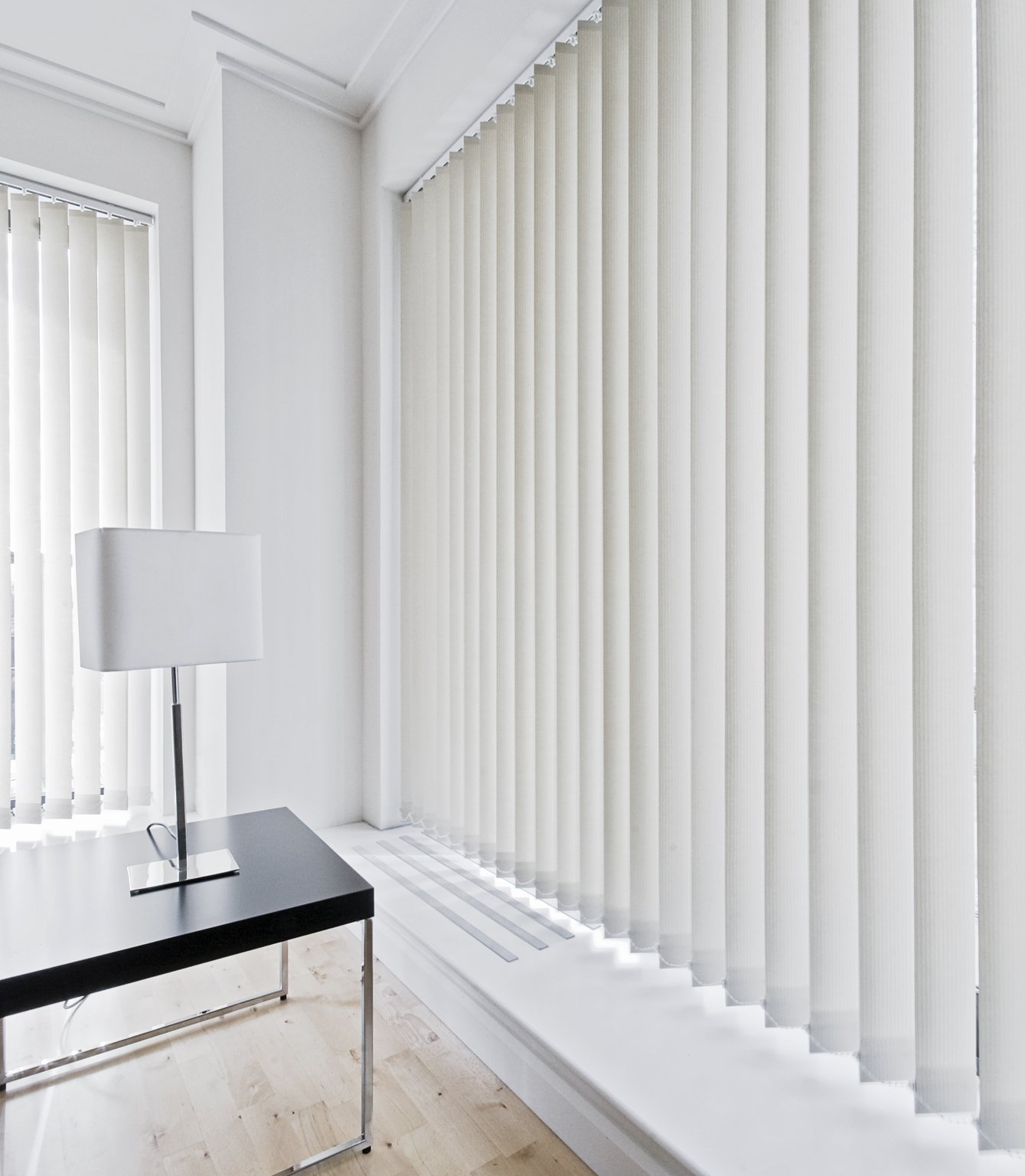 View of vertical blinds on both wall of loungeroom. White lamp on black and chrome coffee table, with light timber flooring.