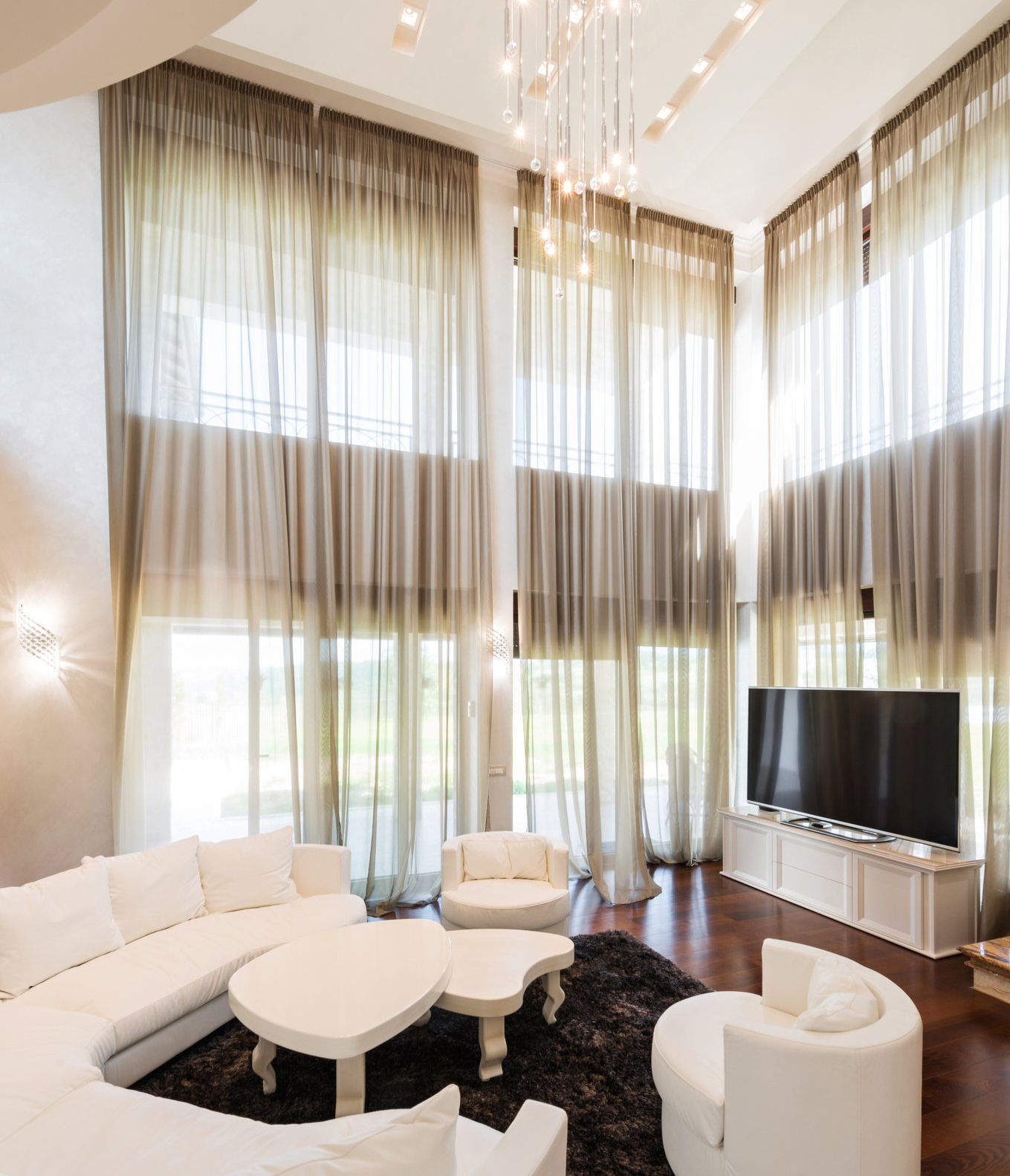 Floor to ceiling sheets curtains in front of roller blinds pulled half way up, in lounge with stark white loungesuite and coffee tables on dark rug. TV on white cabinet against one window wall.