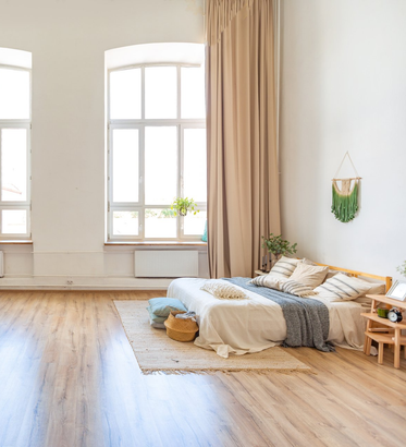 Bedroom with timber floor and bed on top of a rug. Floor to ceiling curtains opened over window with light flowing through.