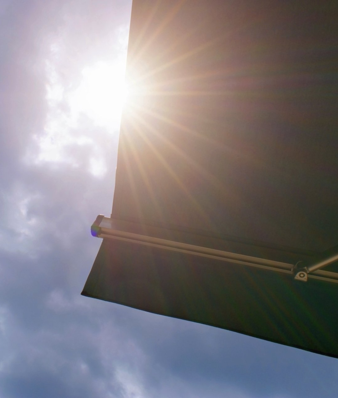 Close up of corner of black canvas extendable awning, showing sun shining and blue sky above.