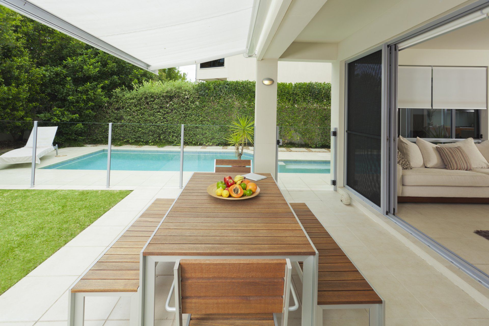 Awning extended over outdoor entertainment area with timber table and seats, with pool in background and green grass in foreground.