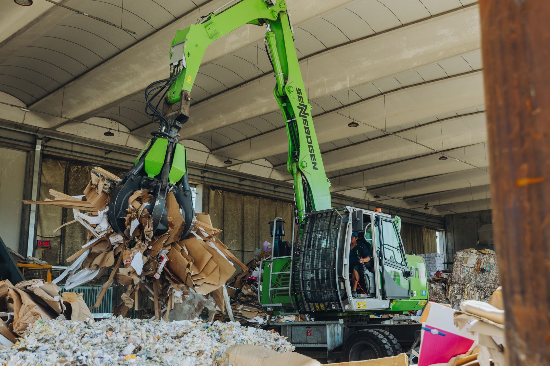 Un escavatore verde frantuma detriti all'interno di un magazzino o di un cantiere di demolizione, con cumuli di macerie tutt'intorno.
